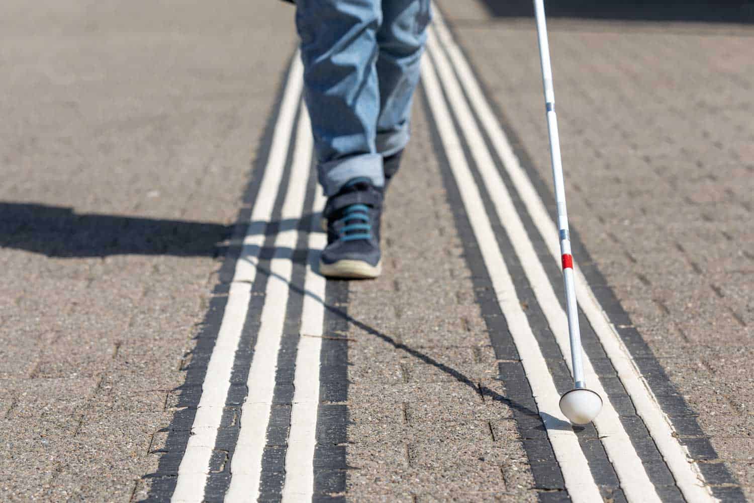 Beine einer sehbeeinträchtigten Person mit Blindenstock neben weissen Bodenleitlinien für Blinde. Beine einer sehbeeinträchtigten Person mit Blindenstock neben weissen Bodenleitlinien für Blinde.
