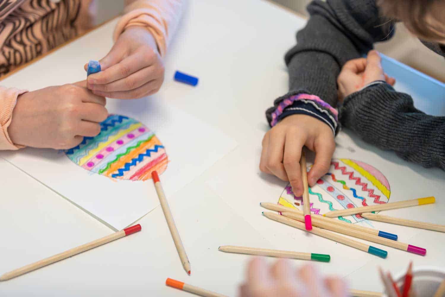 Jugendliche malen Eier auf Papier mit bunten Stiften auf einem Tisch.