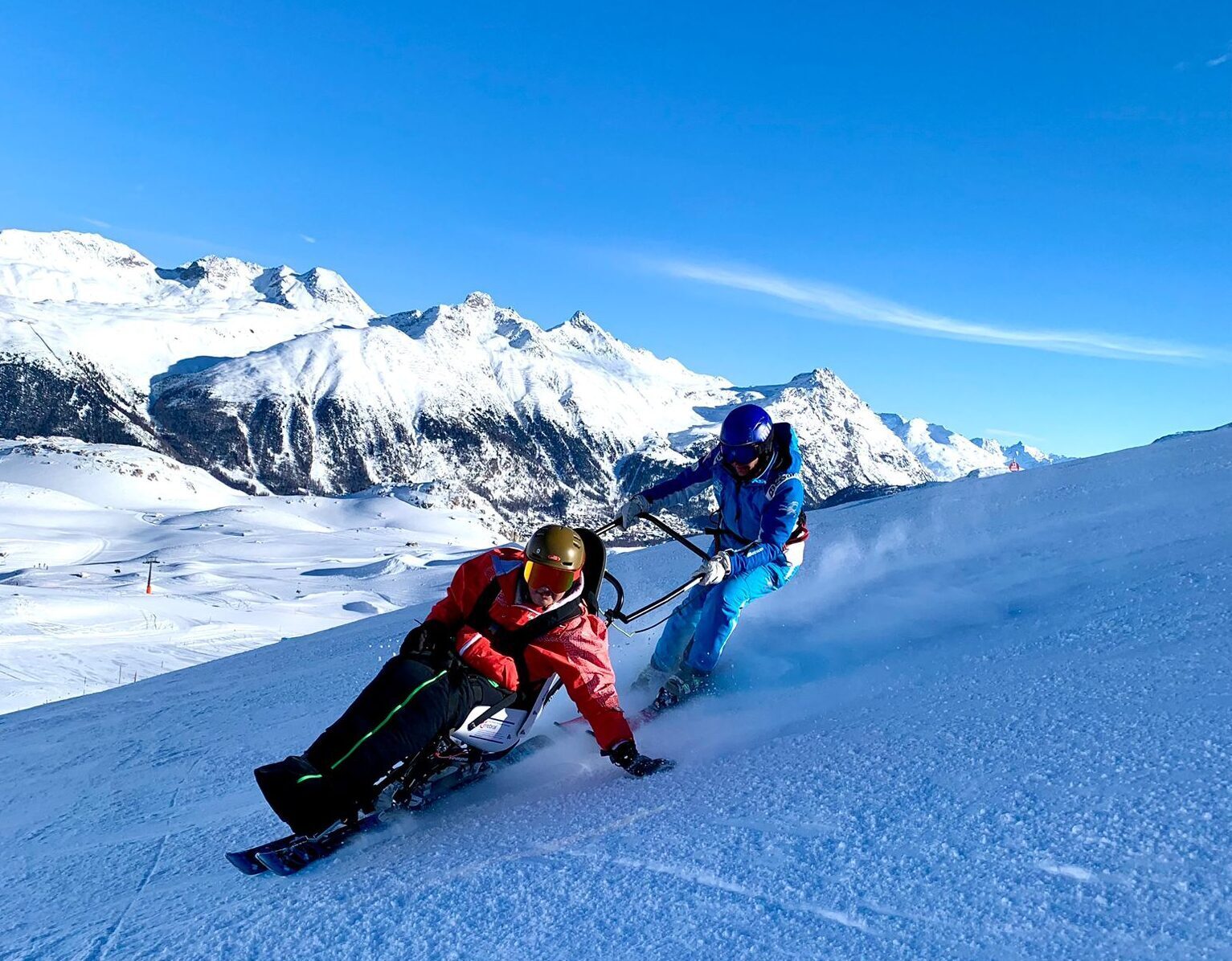 Schülerin mit Monoski und Skilehrperson auf der Piste. Im Hintergrund ist ein beeindruckendes Bergpanorama zu sehen.