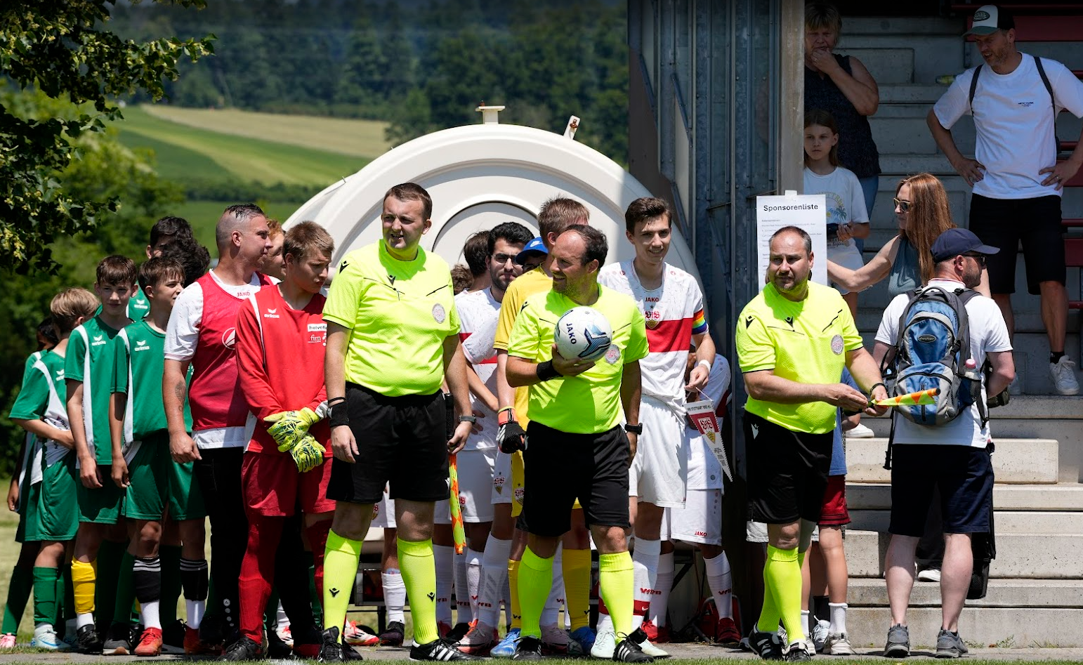 Fussballspiel Fronleichnam Auf dem Foto sind die zwei gemischte Teams – bestehend aus Schülerinnen des Kompetenzzentrums SONNENBERG, Jugendlichen vom FC Bruschgol, dem Team „Brustring der Herzen“ des VfB Stuttgart sowie C-Junioren des FC Baar zu sehen wie sie auflaufen. Alle Spieler*innen sind in farbigen Trikots und voller Freude dabei. Im Hintergrund sitzen und stehen über 100 Zuschauer*innen auf der grossen Tribüne. Am Spielfeldrand beobachten drei Offizielle aufmerksam das Geschehen. Die Szene strahlt Gemeinschaft, Inklusion und sportliche Begeisterung aus.