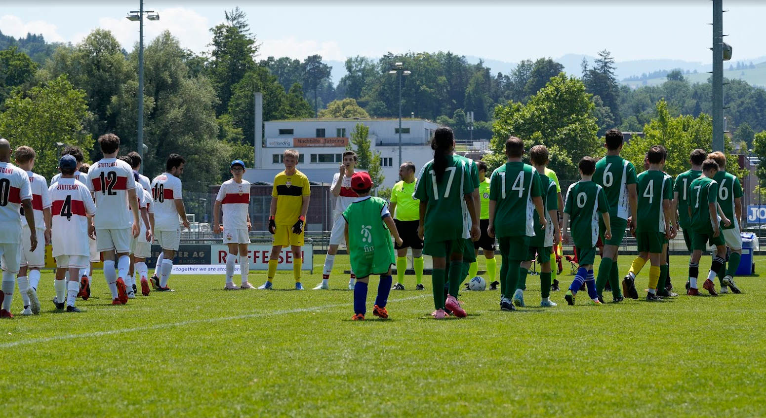 Fussballspiel Fronleichnam_1 Auf dem Foto sind die zwei gemischte Teams – bestehend aus Schülerinnen des Kompetenzzentrums SONNENBERG, Jugendlichen vom FC Bruschgol, dem Team „Brustring der Herzen“ des VfB Stuttgart sowie C-Junioren des FC Baar zu sehen wie sie auflaufen. Alle Spieler*innen sind in farbigen Trikots und voller Freude dabei. Im Hintergrund sitzen und stehen über 100 Zuschauer*innen auf der grossen Tribüne. Am Spielfeldrand beobachten drei Offizielle aufmerksam das Geschehen. Die Szene strahlt Gemeinschaft, Inklusion und sportliche Begeisterung aus.
